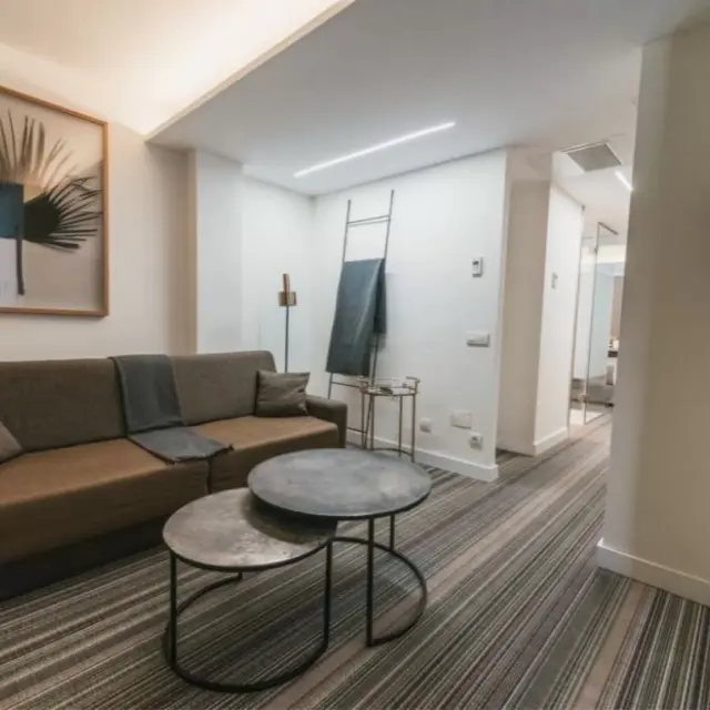 Interior de una suite con sofá marrón de tres plazas, cojines y mantas grises, mesa de centro redonda de dos alturas