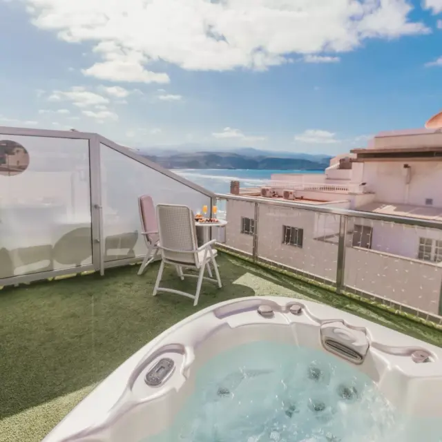 Jacuzzi en terraza privada con césped artificial, dos sillas y mesa, rodeado de barandilla de cristal con vistas