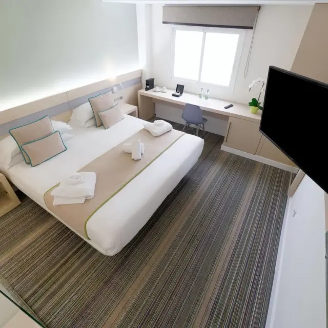 Vista general de una suite de hotel con cama king size vestida de blanco y beige, cojines decorativos, toallas