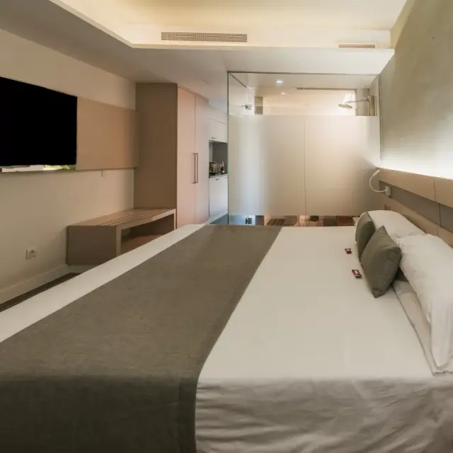 Vista de una suite de hotel con una cama grande vestida de blanco y gris, cabecero iluminado, televisión de pantalla