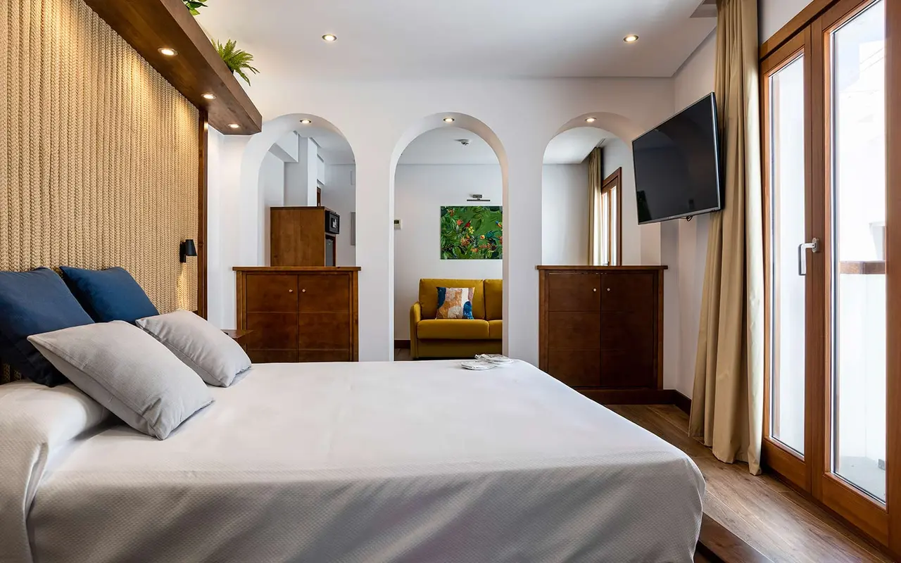 Vista de una habitación junior suite con cama grande vestida de blanco, cabecero de tela, arcos que conectan