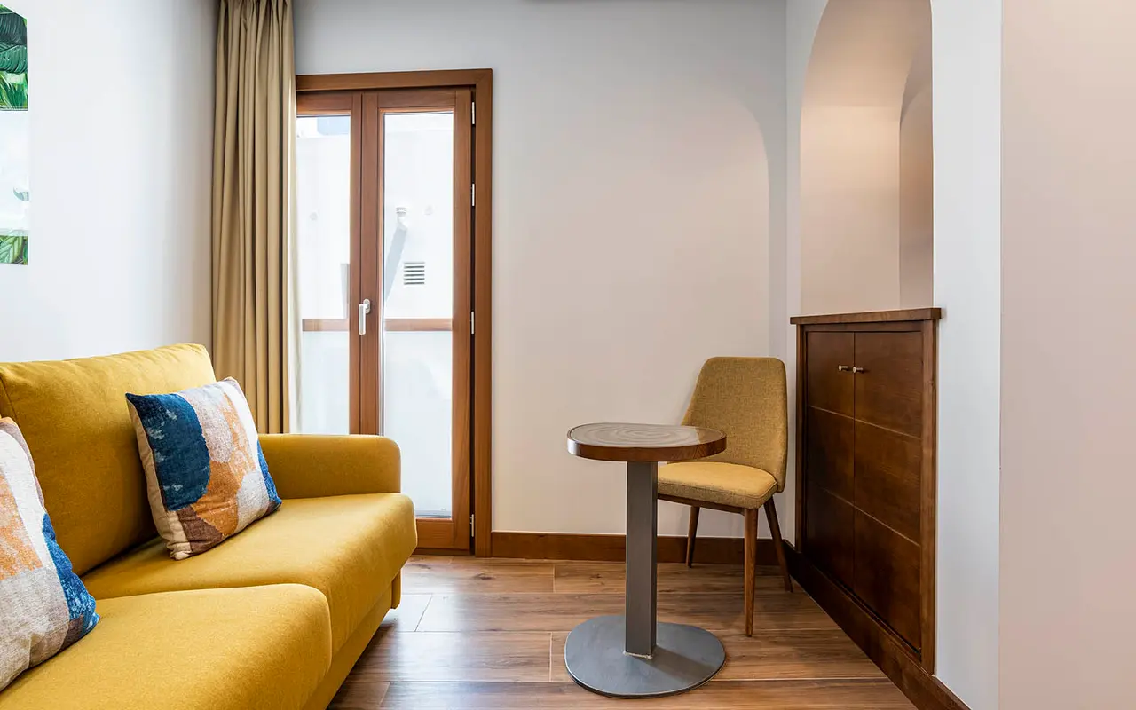 Interior de una habitación junior suite con sofá amarillo de dos plazas, mesa redonda con silla tapizada, armario