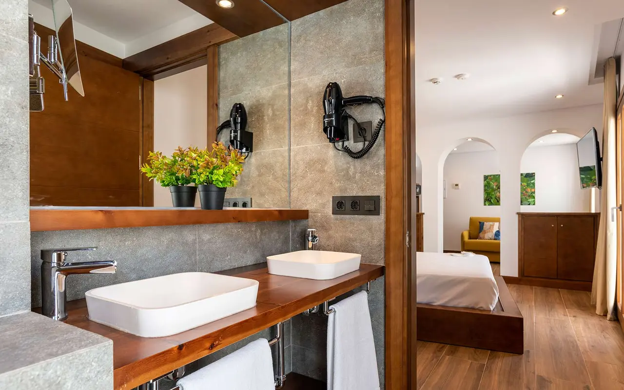 Baño de suite con doble lavabo rectangular blanco sobre encimera de madera, grifería cromada, espejo grande, secadores