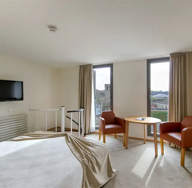 Interior de una suite con cama grande en primer plano, dos sillones de cuero naranja y mesa redonda junto a grandes