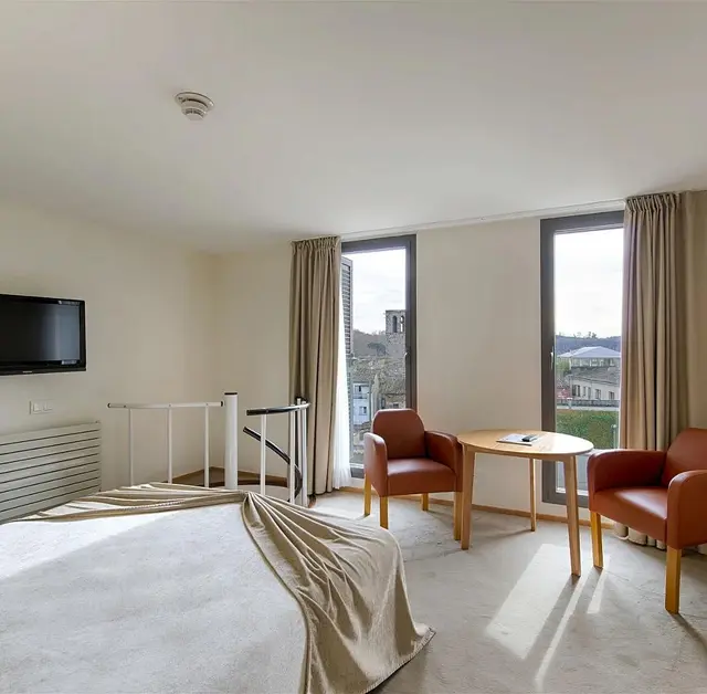 Interior de una suite con cama grande en primer plano, dos sillones de cuero naranja y mesa redonda junto a grandes