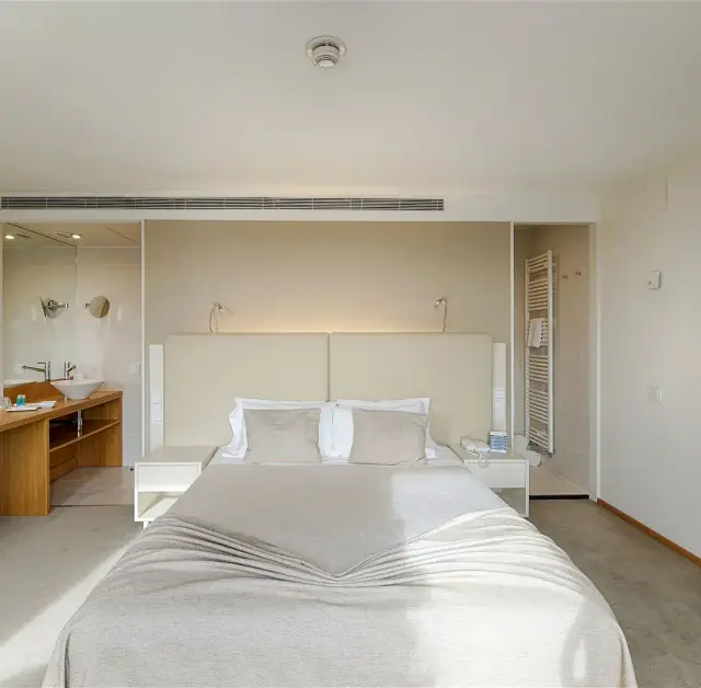 Vista de una suite de hotel con una cama grande en el centro, cabecero beige, mesitas de noche blancas, un escritorio