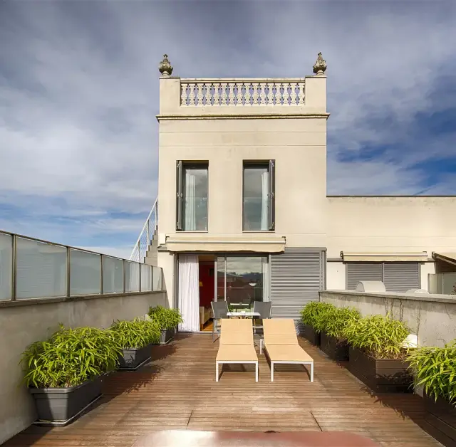 Terraza exterior con suelo de madera, dos tumbonas, macetas con plantas y vistas a un edificio de estilo clásico