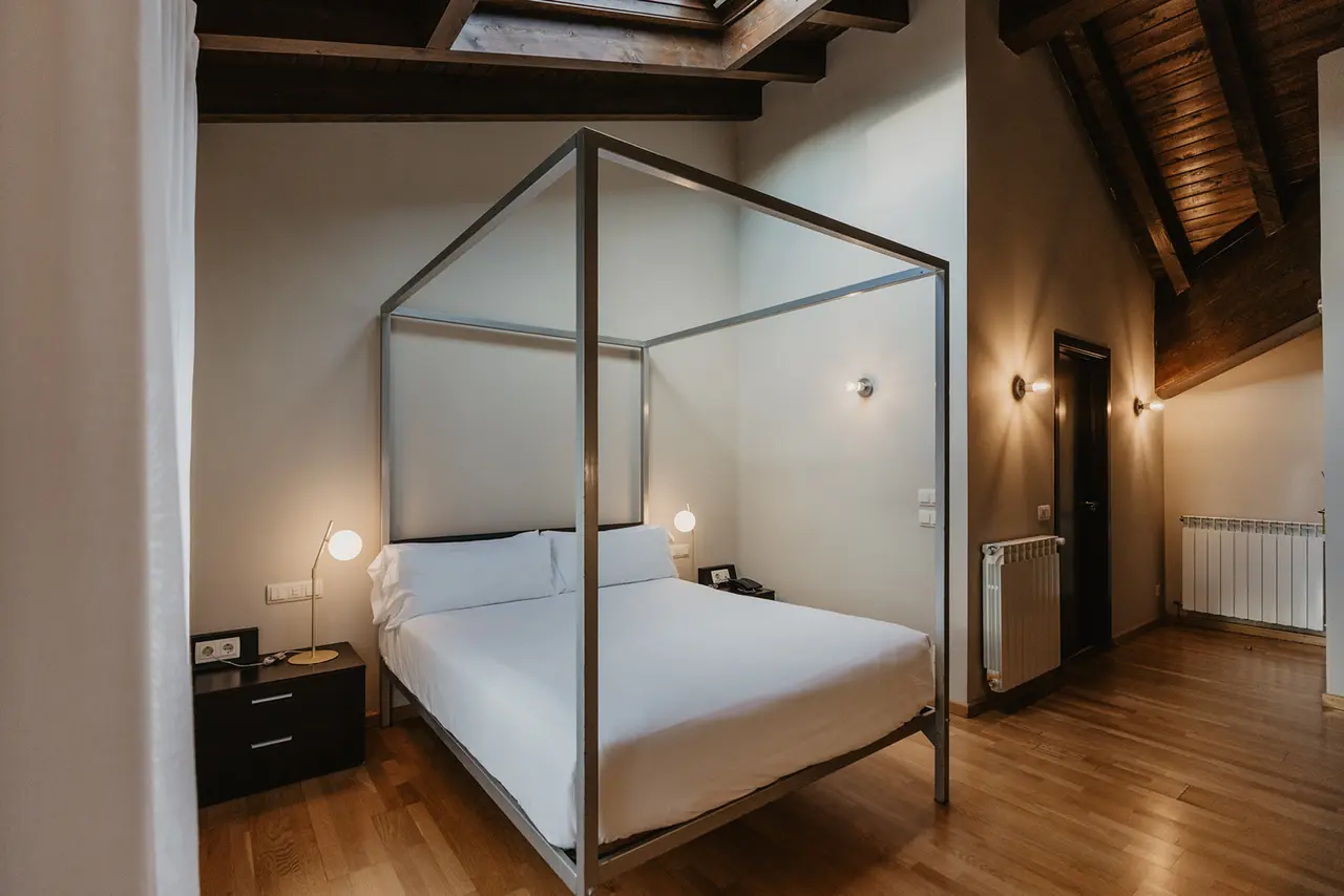 Habitación con cama de matrimonio de cuatro postes con dosel metálico, sábanas blancas, mesitas de noche con lámparas