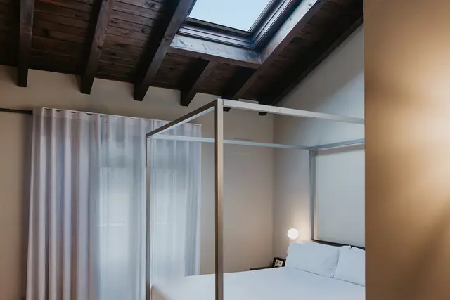Habitación con cama de dosel metálico, dosel con cortinas blancas, techo de vigas de madera oscura y claraboya