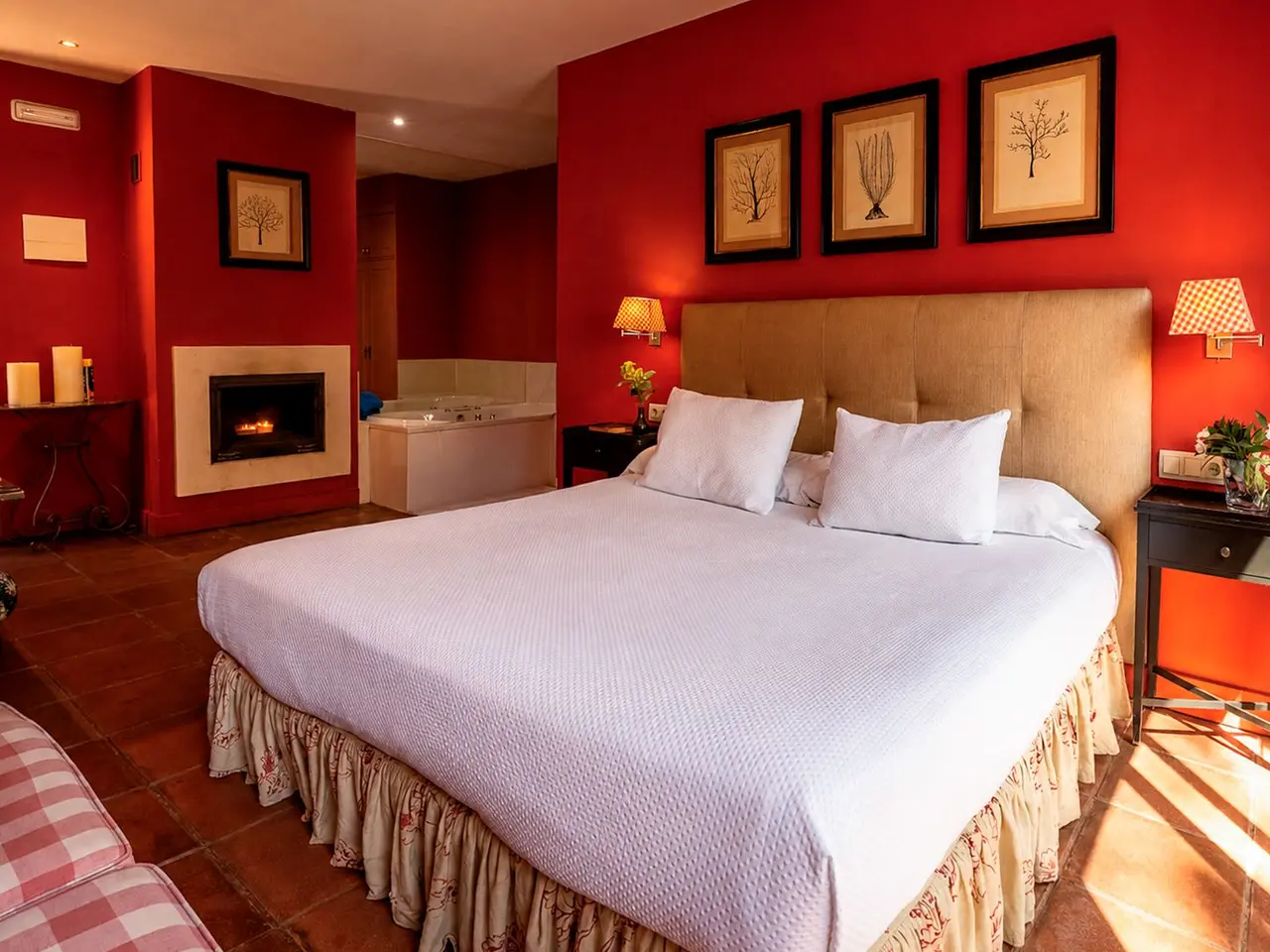 Interior de una suite con paredes rojas, una gran cama con dosel beige y sábanas blancas, chimenea encendida al fondo
