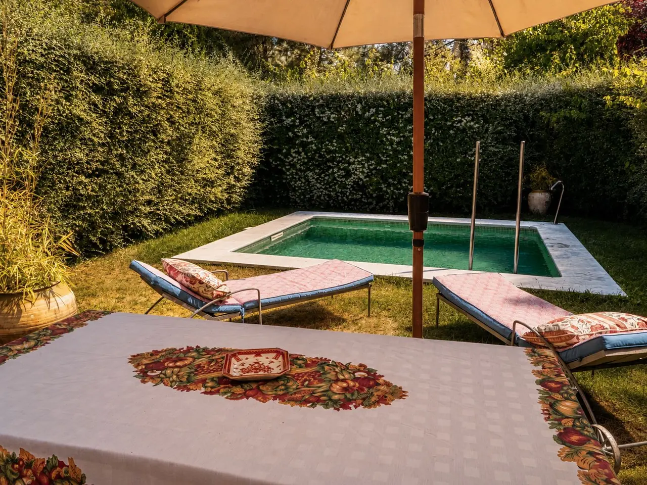 Piscina rectangular privada rodeada de césped, con dos tumbonas, una mesa con mantel en primer plano y una sombrilla.