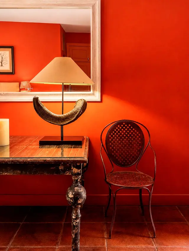 Interior de habitación con paredes rojas, mesa de madera antigua con lámpara y escultura de cuerno, silla de metal