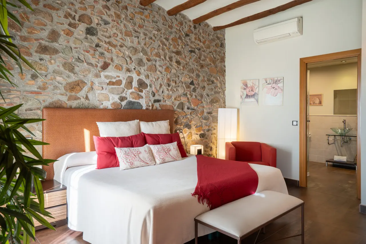Habitación con cama grande vestida en blanco y rojo, cabecero naranja, pared de piedra rústica, banco tapizado al pie