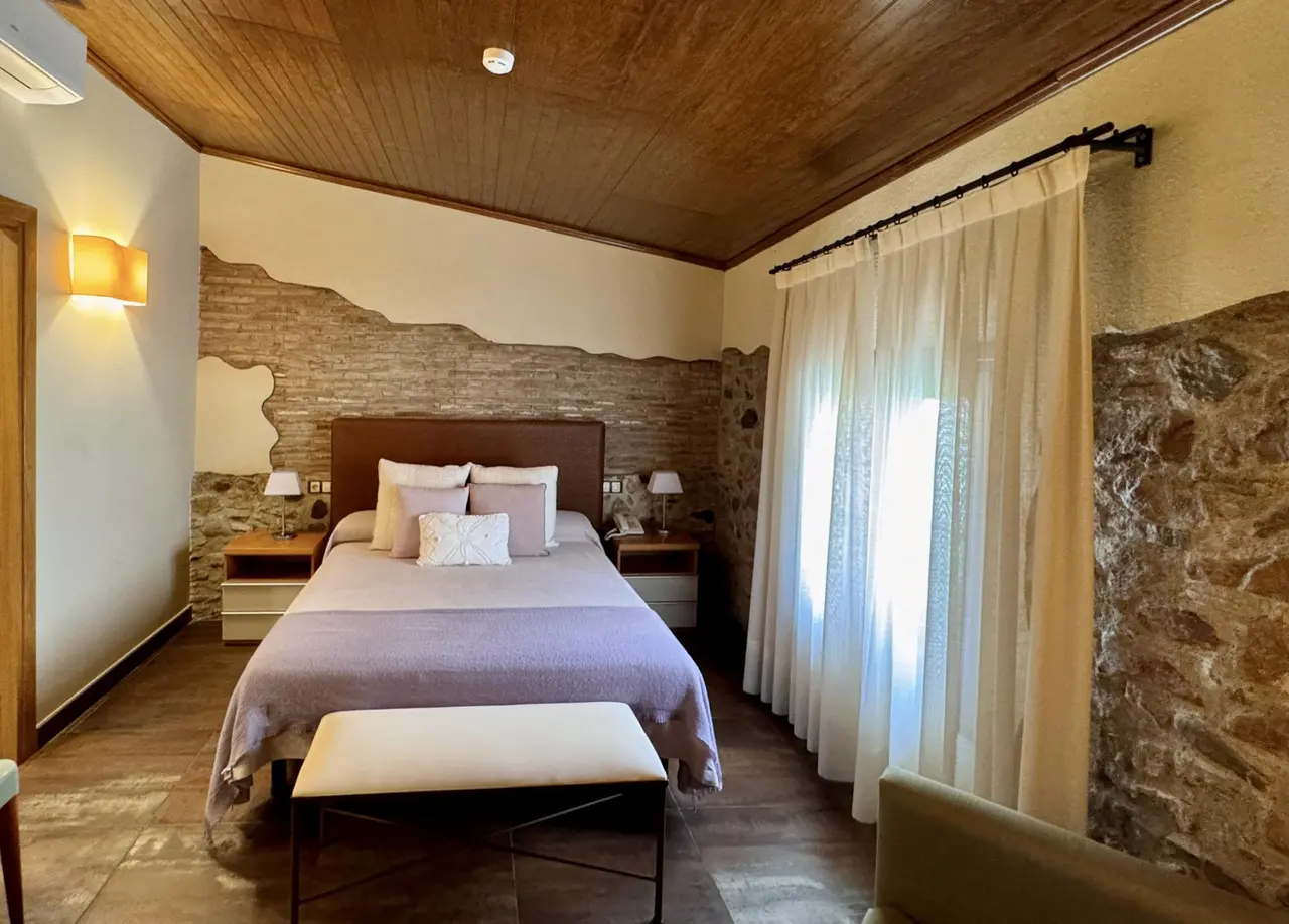Habitación con cama doble, cabecero de madera, mesitas de noche con lámparas, banco tapizado al pie de la cama