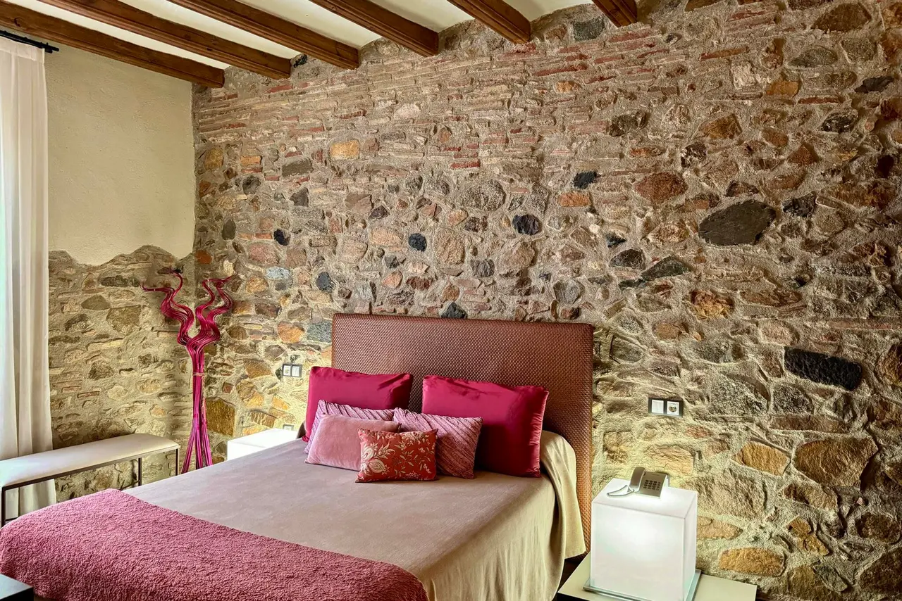 Habitación con cama doble, cabecero textil rosa, cojines en tonos fucsia y rojo, y una pared de piedra rústica.