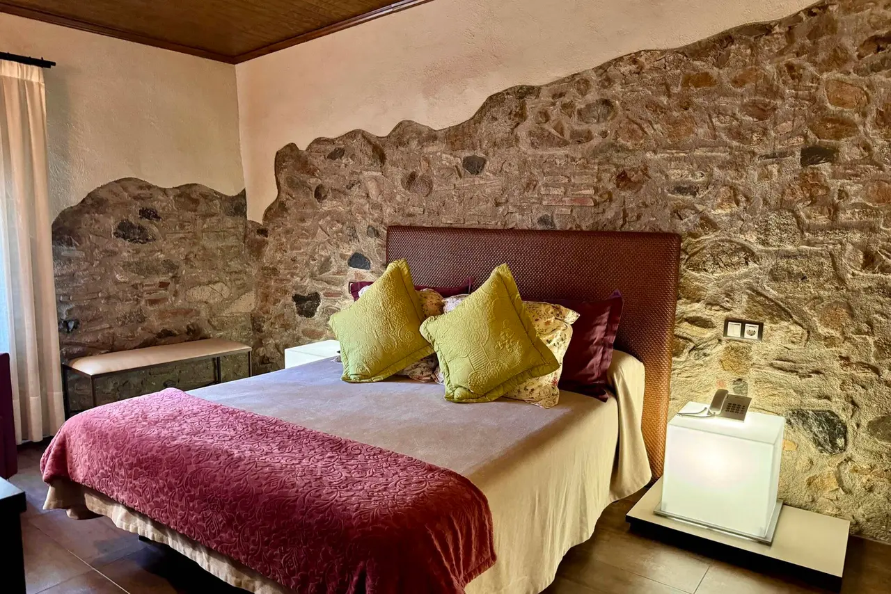 Habitación con cama grande vestida con colcha blanca y manta roja, varios cojines verdes y gran cabecero.