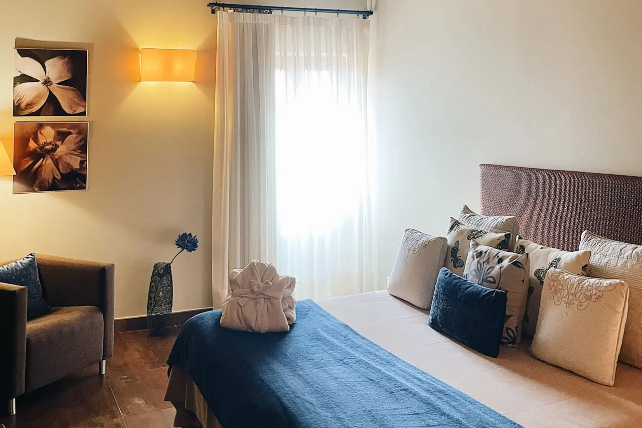 Habitación con cama grande vestida con colcha azul, múltiples cojines decorativos, butaca marrón, ventana con cortinas