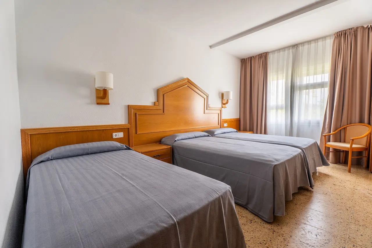Habitación de hotel con dos camas individuales, cabeceros de madera, mesitas de noche, apliques de pared, ventana
