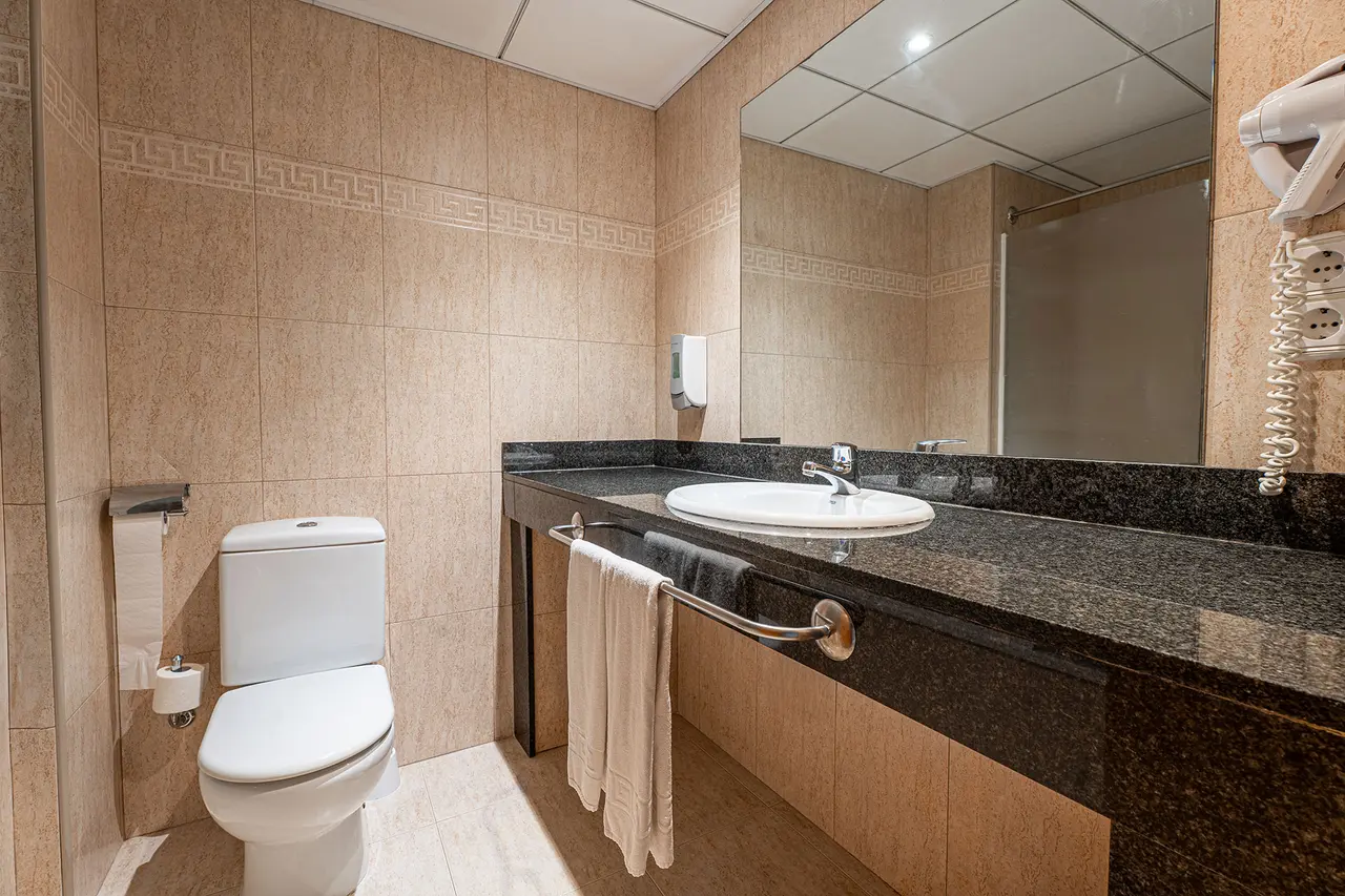Baño de suite con paredes de azulejos beige, inodoro blanco, lavabo redondo sobre encimera de granito negro, espejo