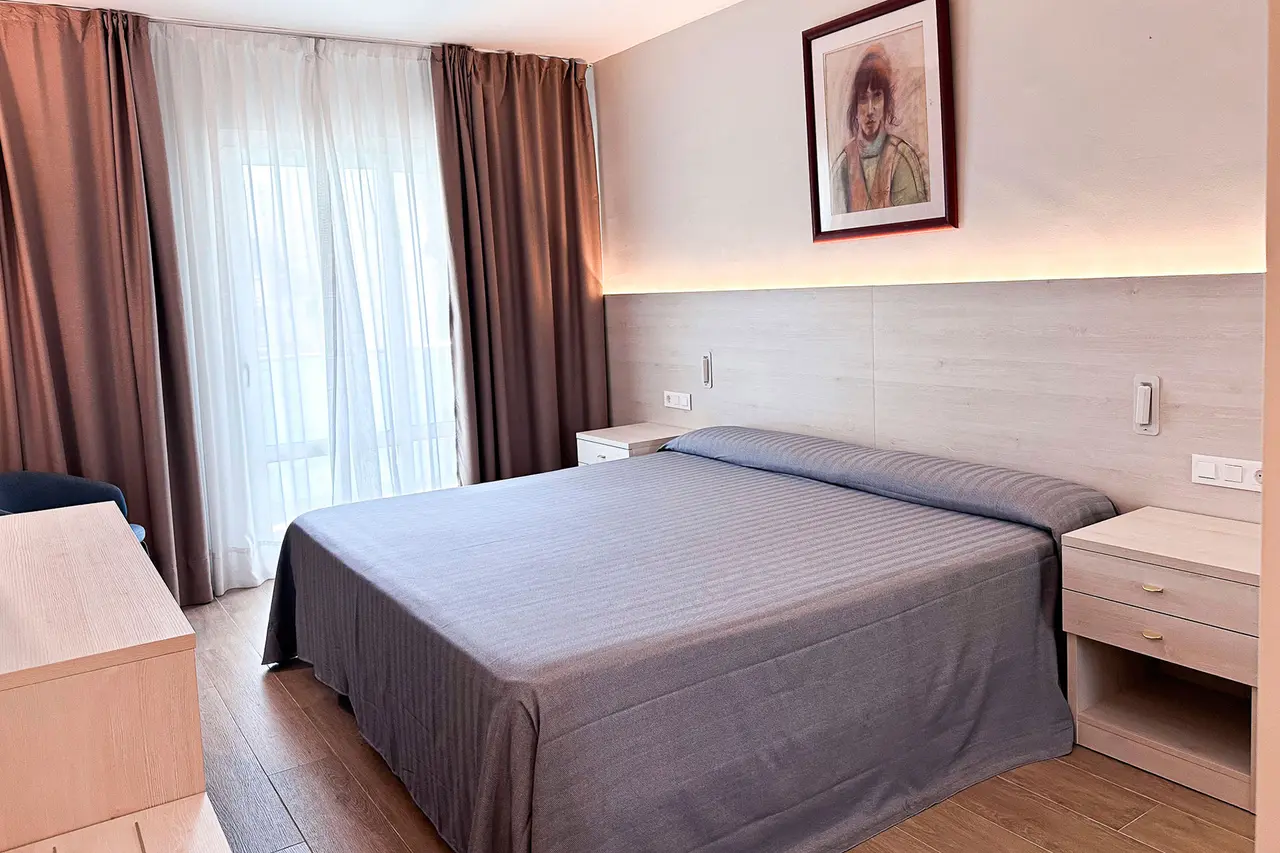 Habitación de hotel con cama doble grande cubierta por un edredón gris, cabecero de madera clara iluminado