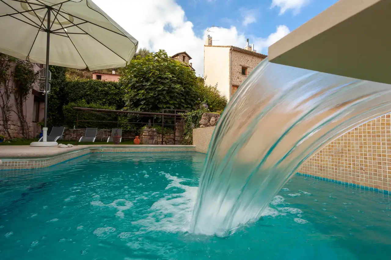 Vista de una piscina exterior con agua turquesa, cascada de agua cayendo desde un borde elevado y un parasol blanco