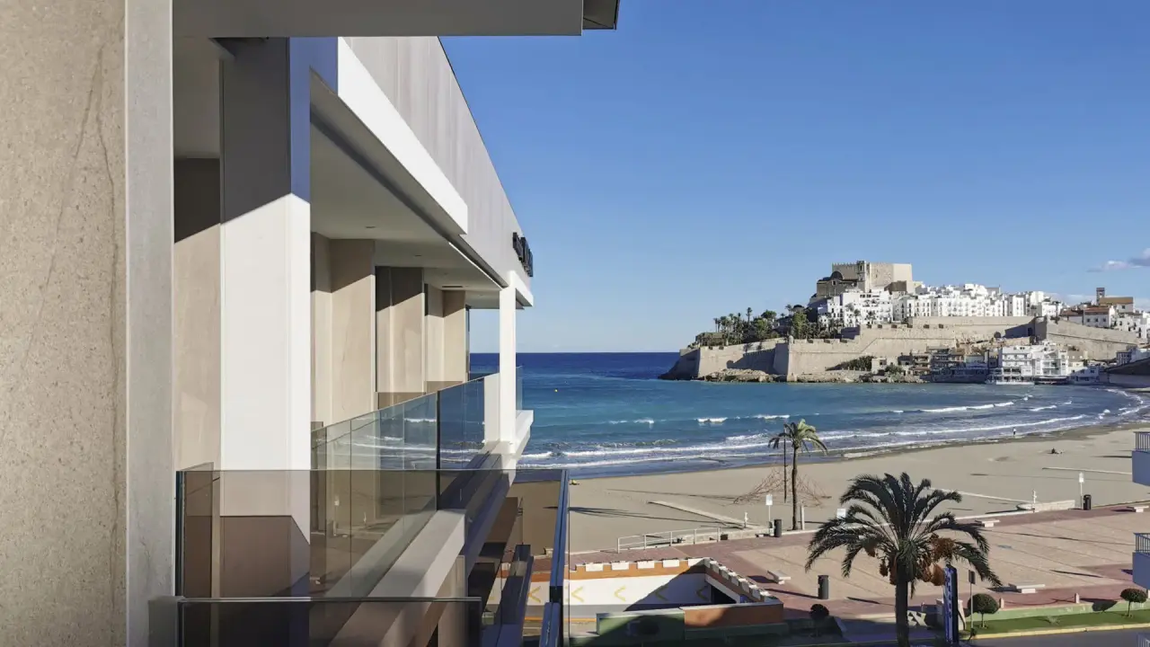 Vista desde una terraza con barandilla de cristal hacia la playa, el mar y el castillo de Peñíscola en el horizonte