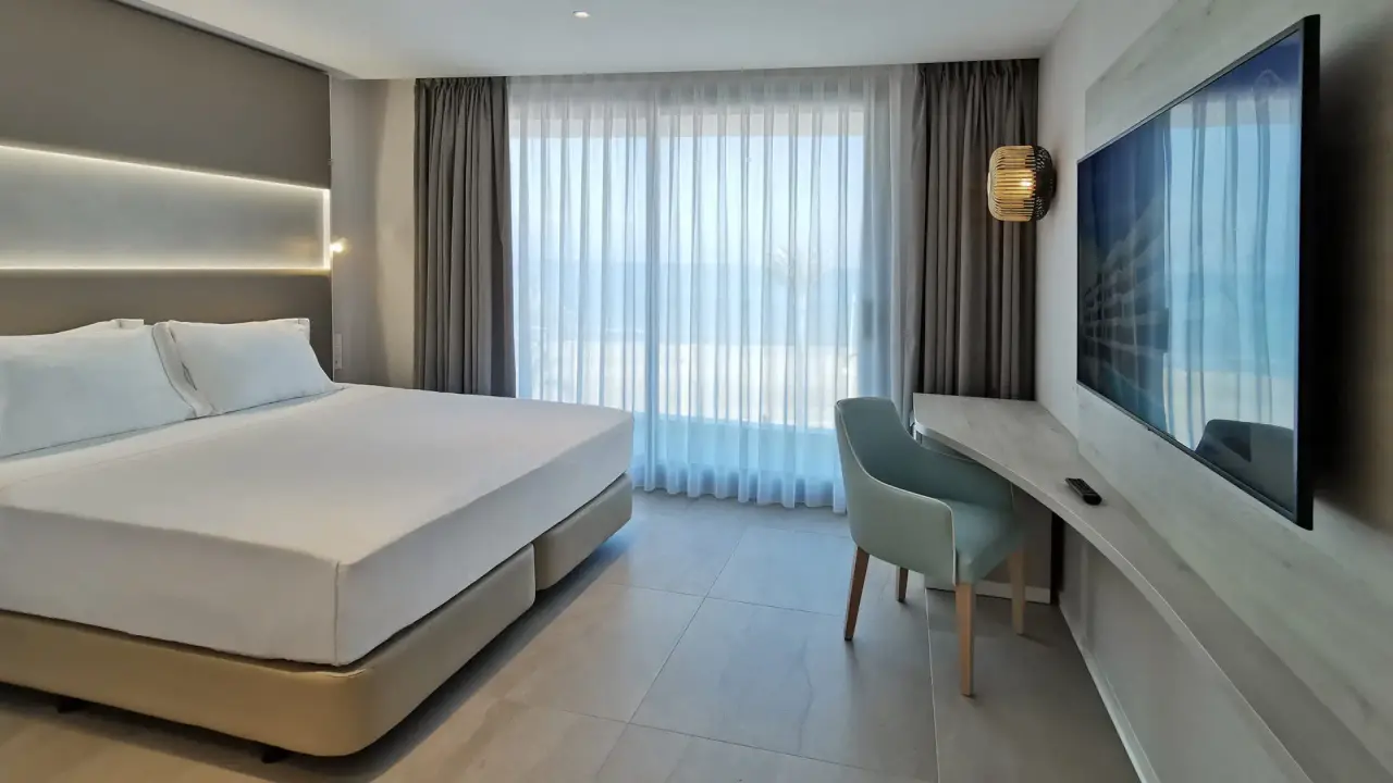 Habitación de hotel con cama doble grande vestida de blanco, escritorio con silla azulada, televisor de pantalla plana