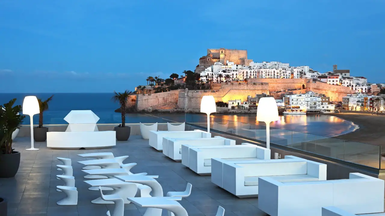 Terraza exterior al atardecer con sillones y tumbonas blancas modernas, lámparas de pie, macetas con plantas y vistas