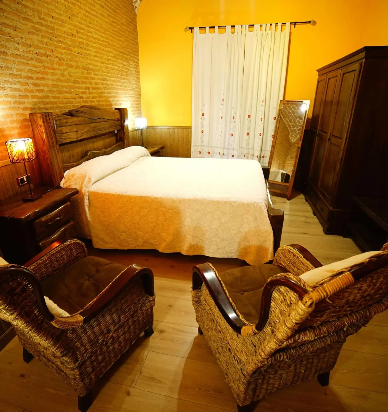 Vista de una suite con cama doble, dos sillones de mimbre en primer plano, mesita de noche con lámpara, armario