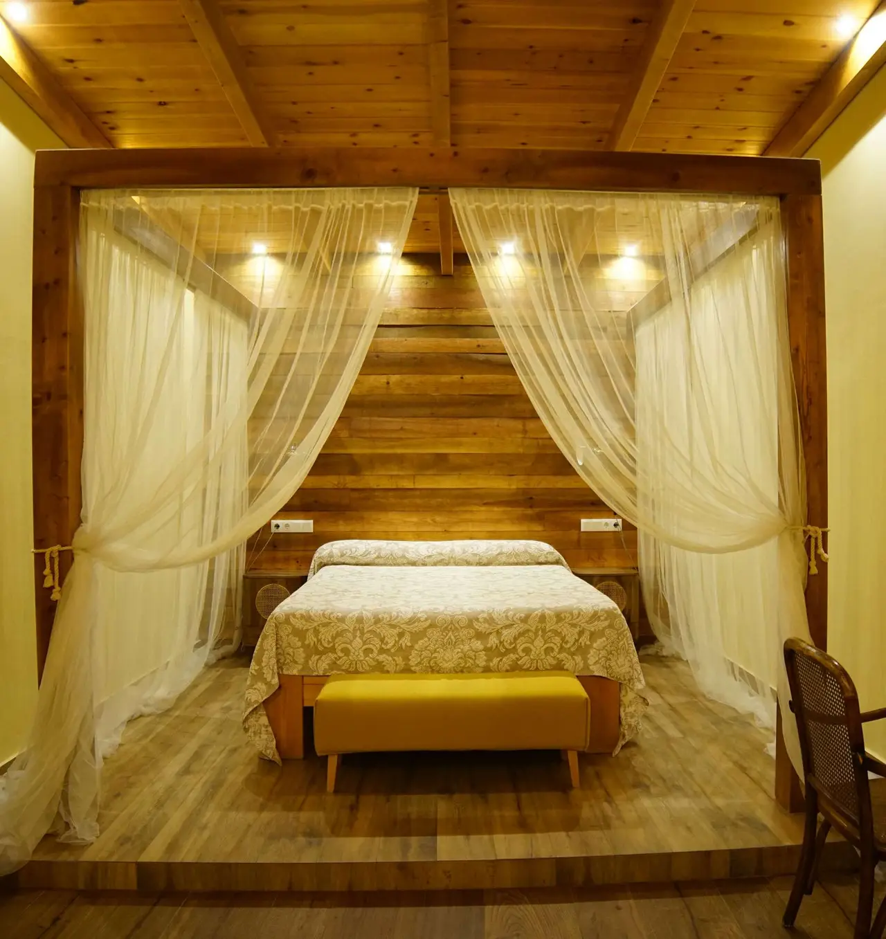 Vista de una suite con cama doble bajo un dosel de madera y cortinas blancas translúcidas, cabecero de pared de madera