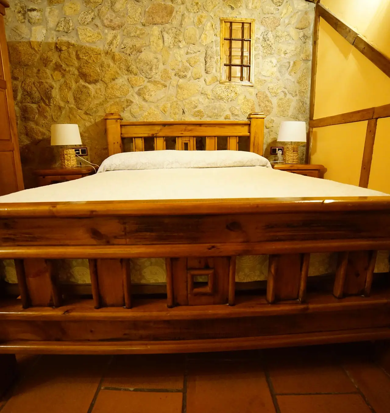 Habitación con cama doble de madera maciza, cabecero alto, mesitas de noche con lámparas y fondo de pared de piedra