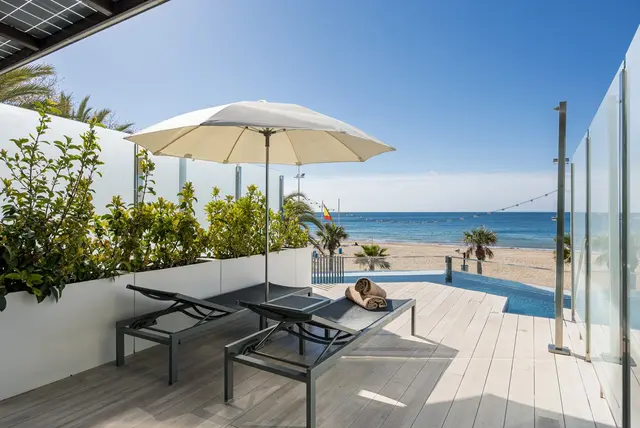 Terraza con piscina privada, dos tumbonas, sombrilla y vistas al mar y la playa.