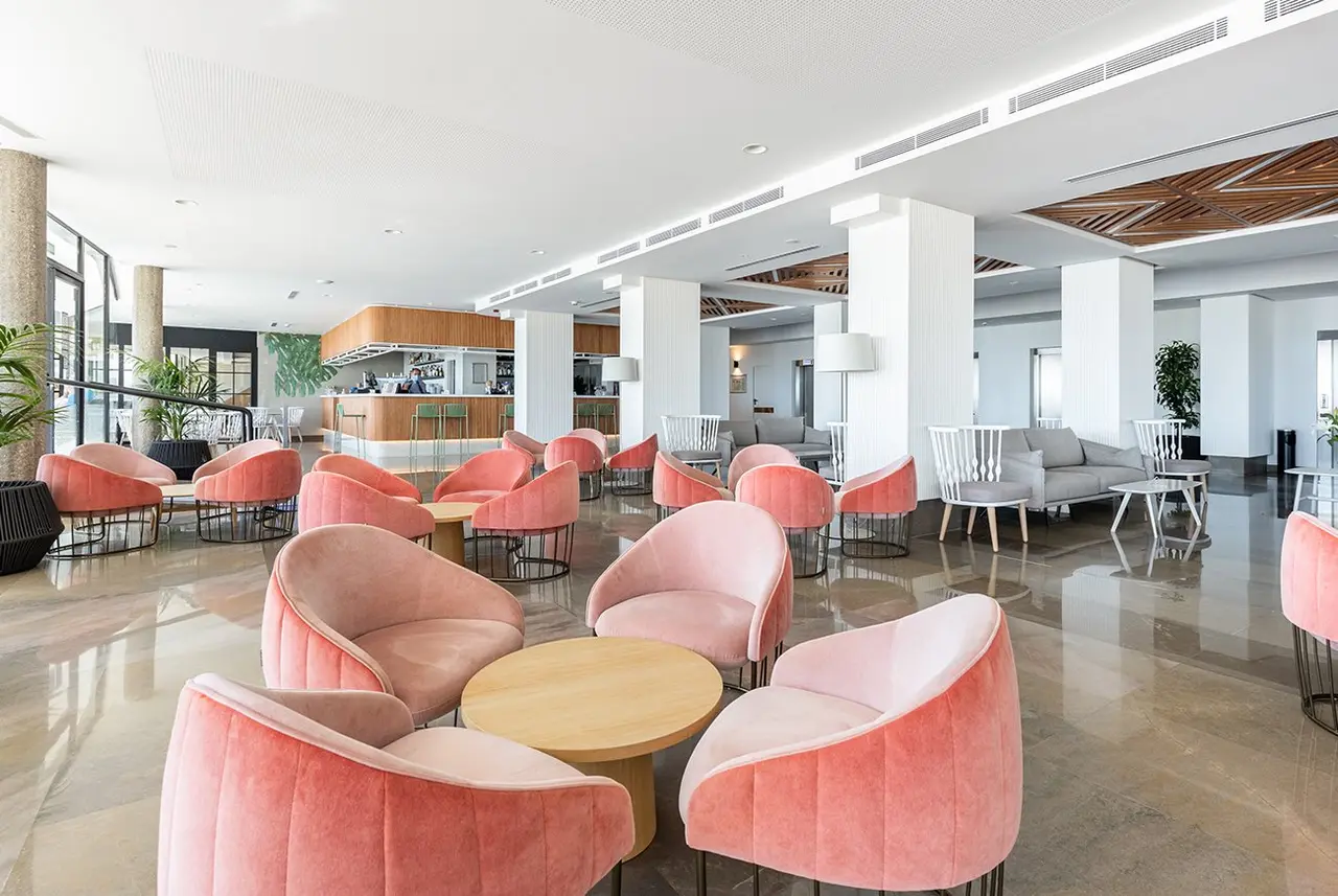 Amplio salón de hotel con múltiples sillones y sofás de color rosa y blanco, mesas de centro redondas de madera, suelo