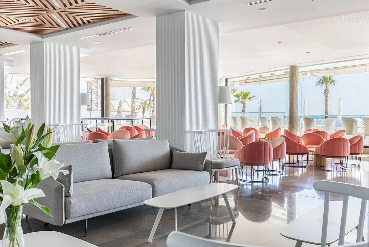 Interior de una sala de estar luminosa con sofá gris en primer plano, mesas blancas, sillas y sillones naranjas