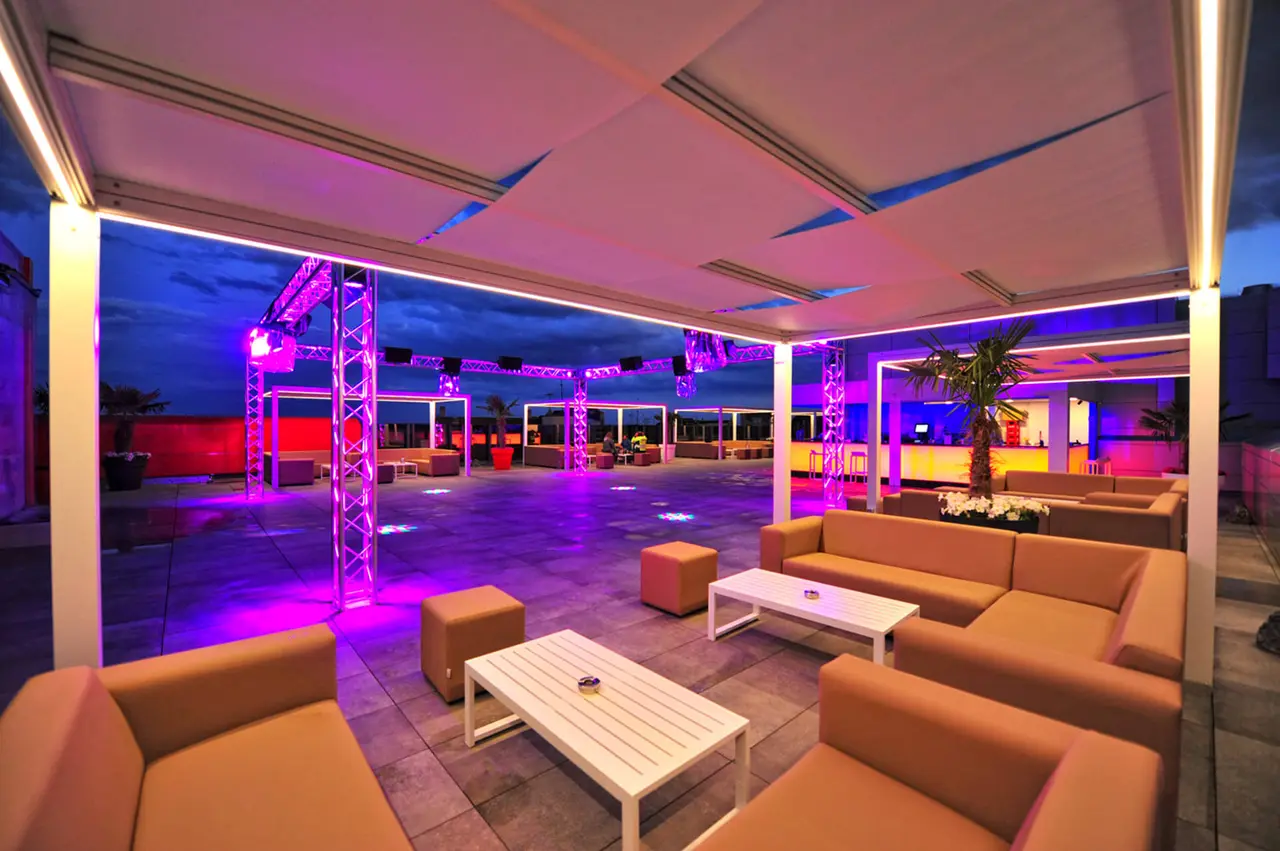 Terraza exterior de hotel con sofás naranjas, mesas de centro blancas y suelo de baldosa, iluminada con luces LED