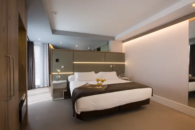 Vista de una suite de hotel con una cama king size vestida de blanco y marrón, cabecero iluminado, mesitas de noche