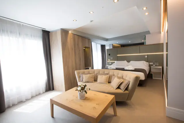 Vista de una suite de hotel con una cama grande al fondo, un sofá beige en el centro, una mesa de centro de madera