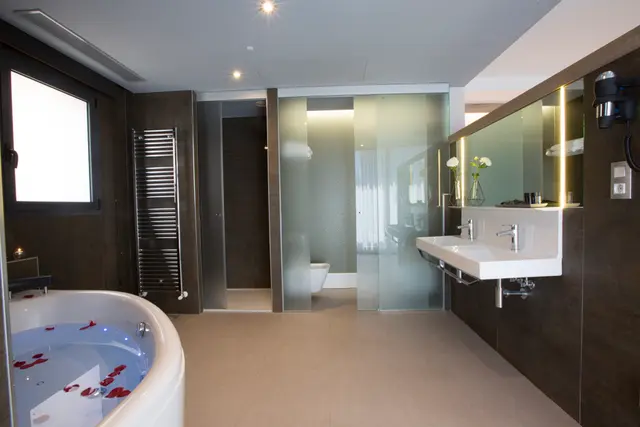 Baño de suite con bañera de hidromasaje blanca en primer plano, llena de agua y pétalos rojos.
