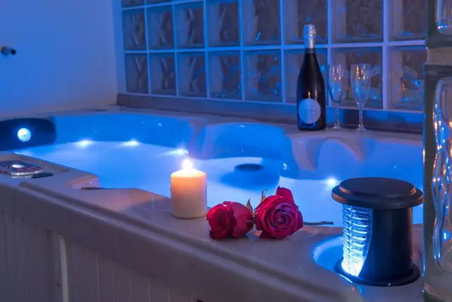 Jacuzzi iluminado en azul con agua, borde de cerámica blanca, velas encendidas, dos rosas rojas y una botella de cava