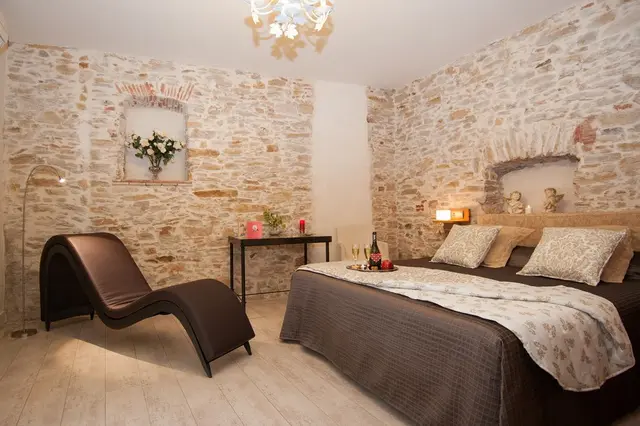 Habitación con paredes de piedra vista, cama doble con almohadas y colcha, chaise longue de cuero marrón, mesa
