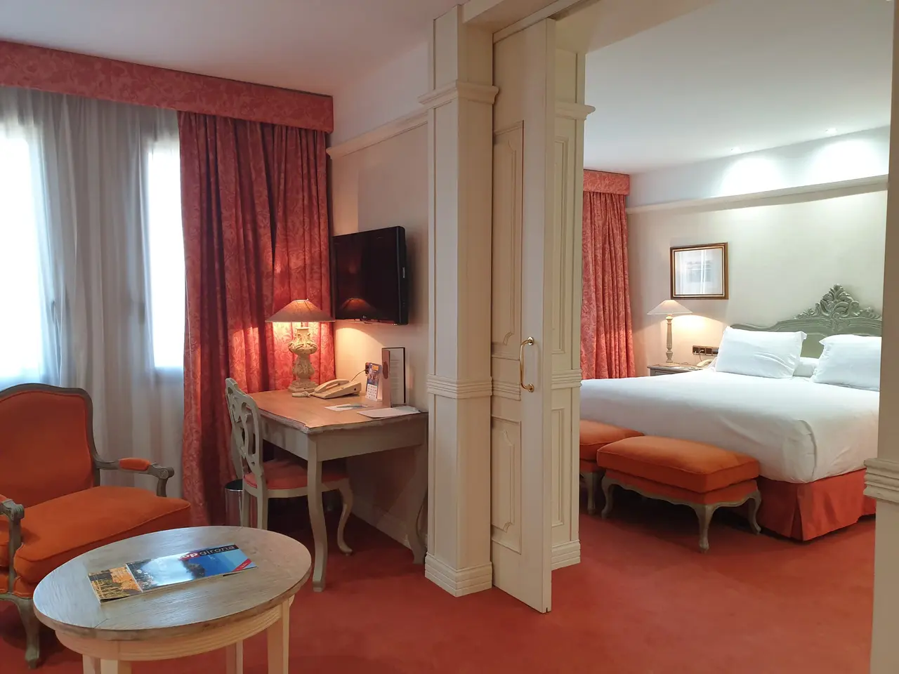 Vista de una suite de hotel con zona de estar que incluye un sillón naranja, una mesa redonda de madera