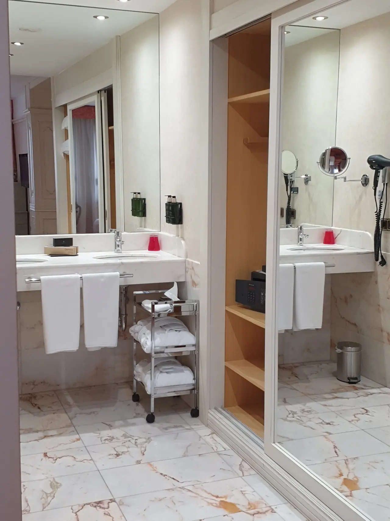 Baño de suite con doble lavabo sobre encimera de mármol, espejo grande, toallas blancas colgadas, carrito auxiliar