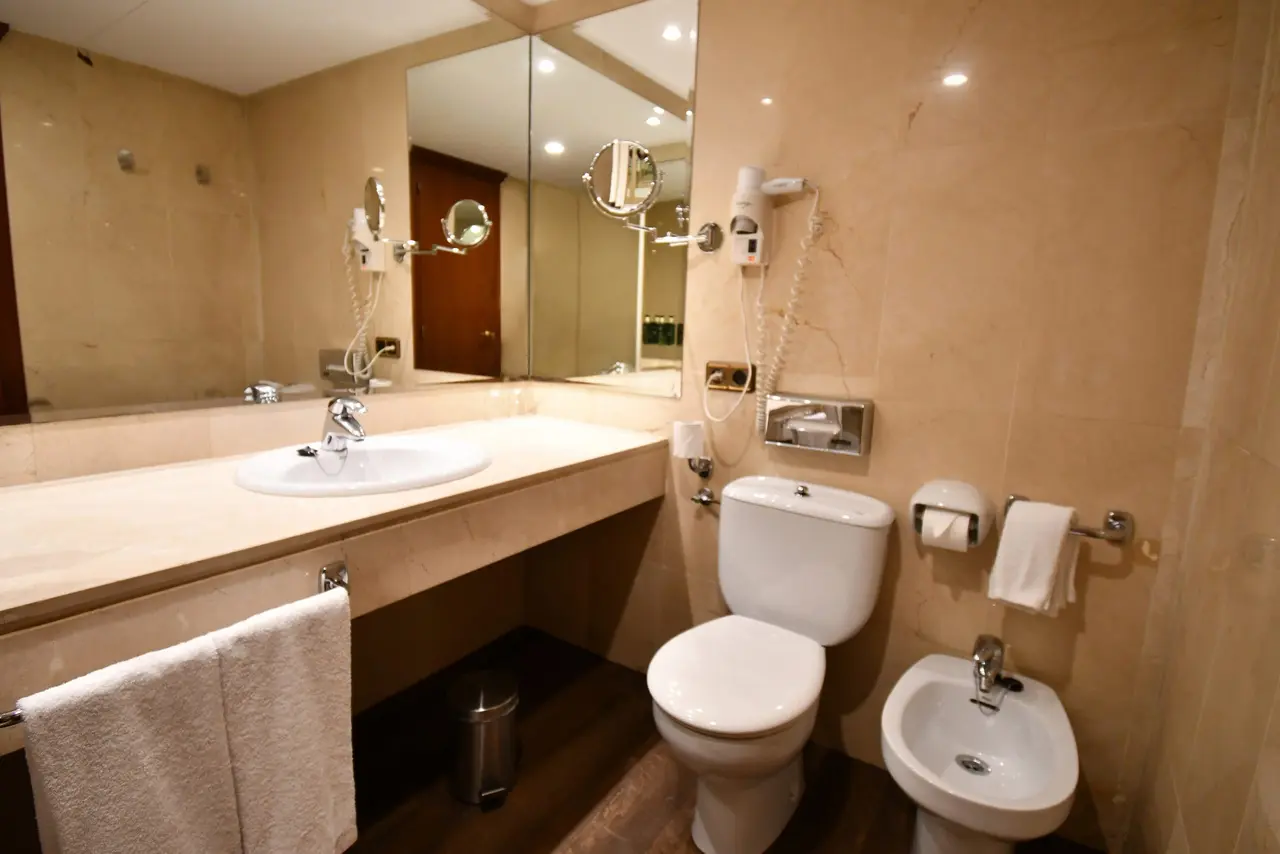 Baño de suite con lavabo sobre encimera de mármol, espejo grande, inodoro, bidé y toallas blancas colgadas.