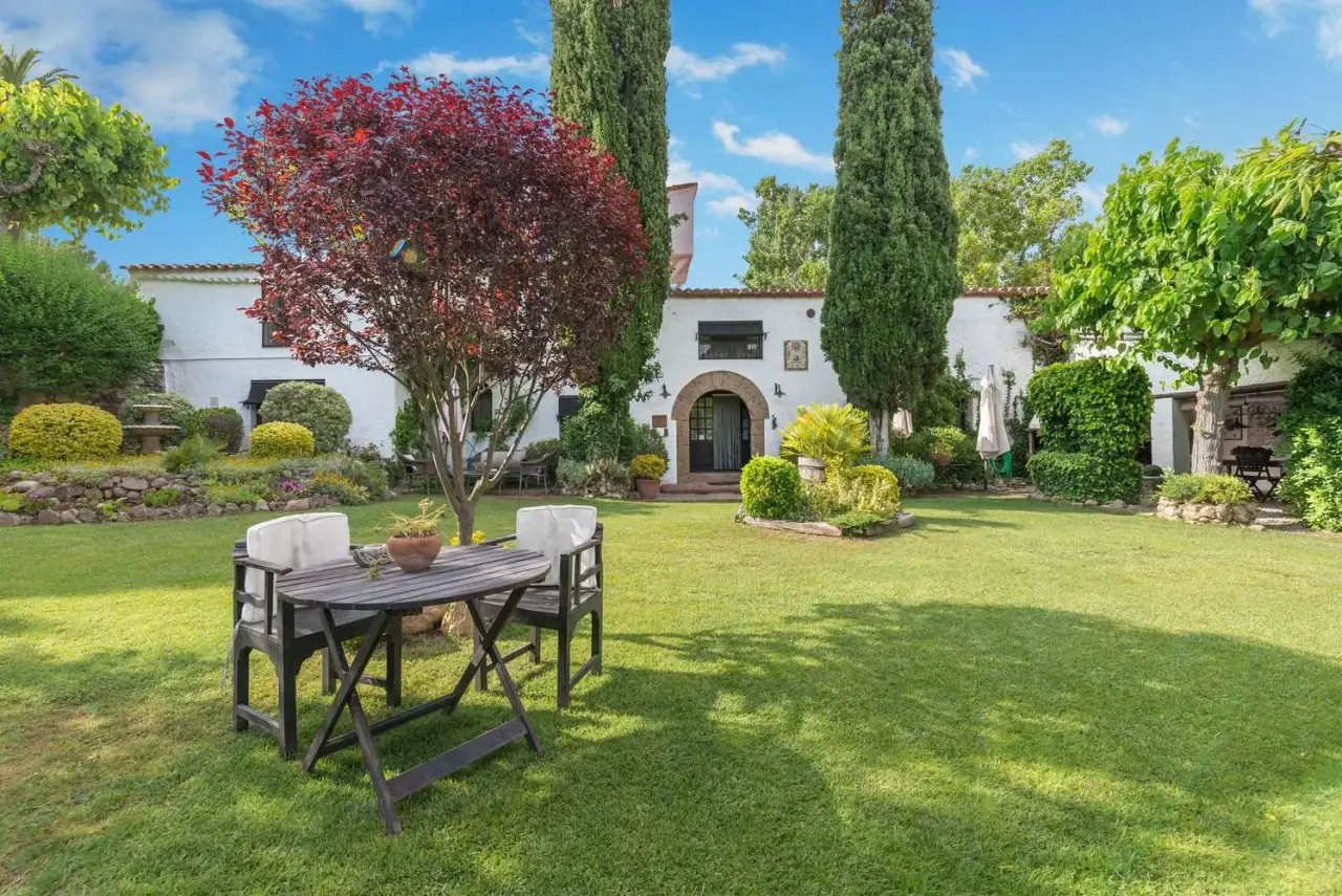 Jardín exterior de una casa blanca con césped verde, mesa y sillas de madera en primer plano, árboles y arbustos