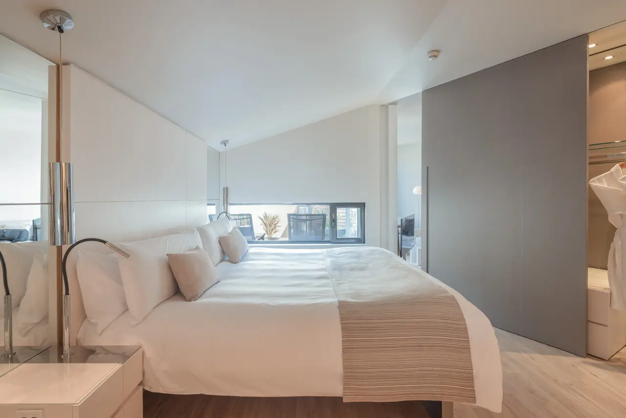 Vista de una suite de hotel con cama king size vestida de blanco, cabecero alto, mesitas de noche modernas