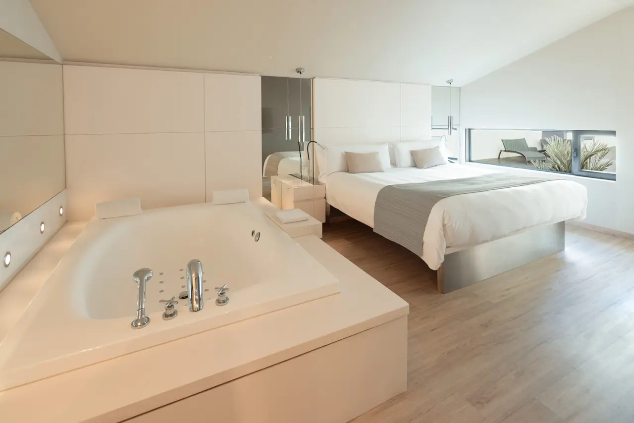 Suite de hotel con una gran bañera de hidromasaje blanca en primer plano, equipada con grifos cromados y jets.