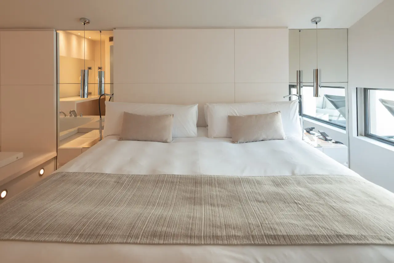 Vista de una suite de hotel con una cama king size vestida con sábanas blancas, cojines beige y un plaid textil