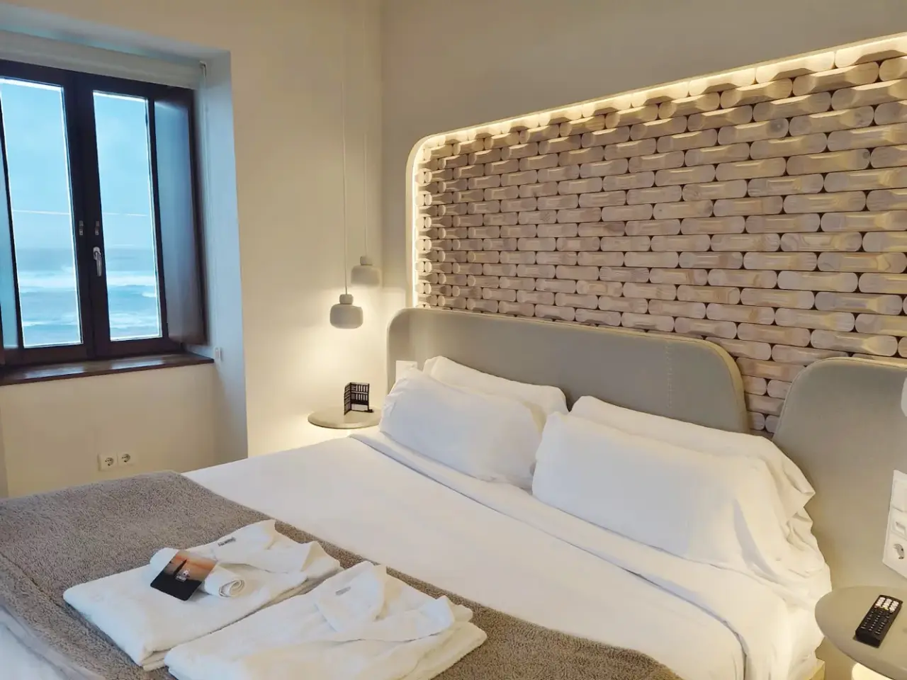 Habitación de hotel con cama grande vestida de blanco, cabecero de obra con pared de ladrillo visto iluminada, ventana