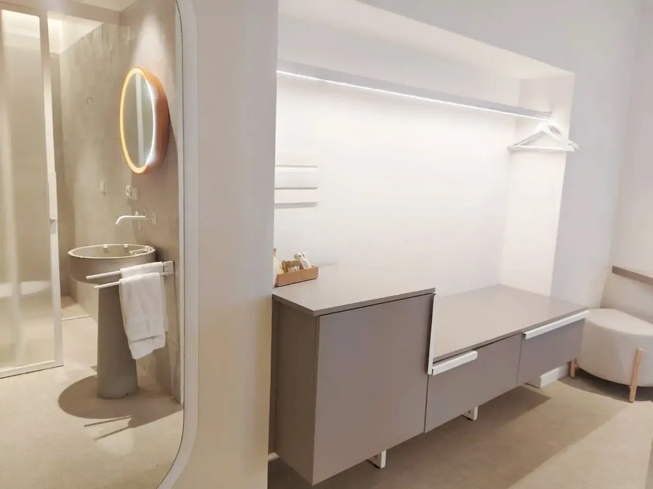 Vista de un baño moderno con lavabo de pedestal, espejo redondo iluminado, toallas blancas y zona de armarios