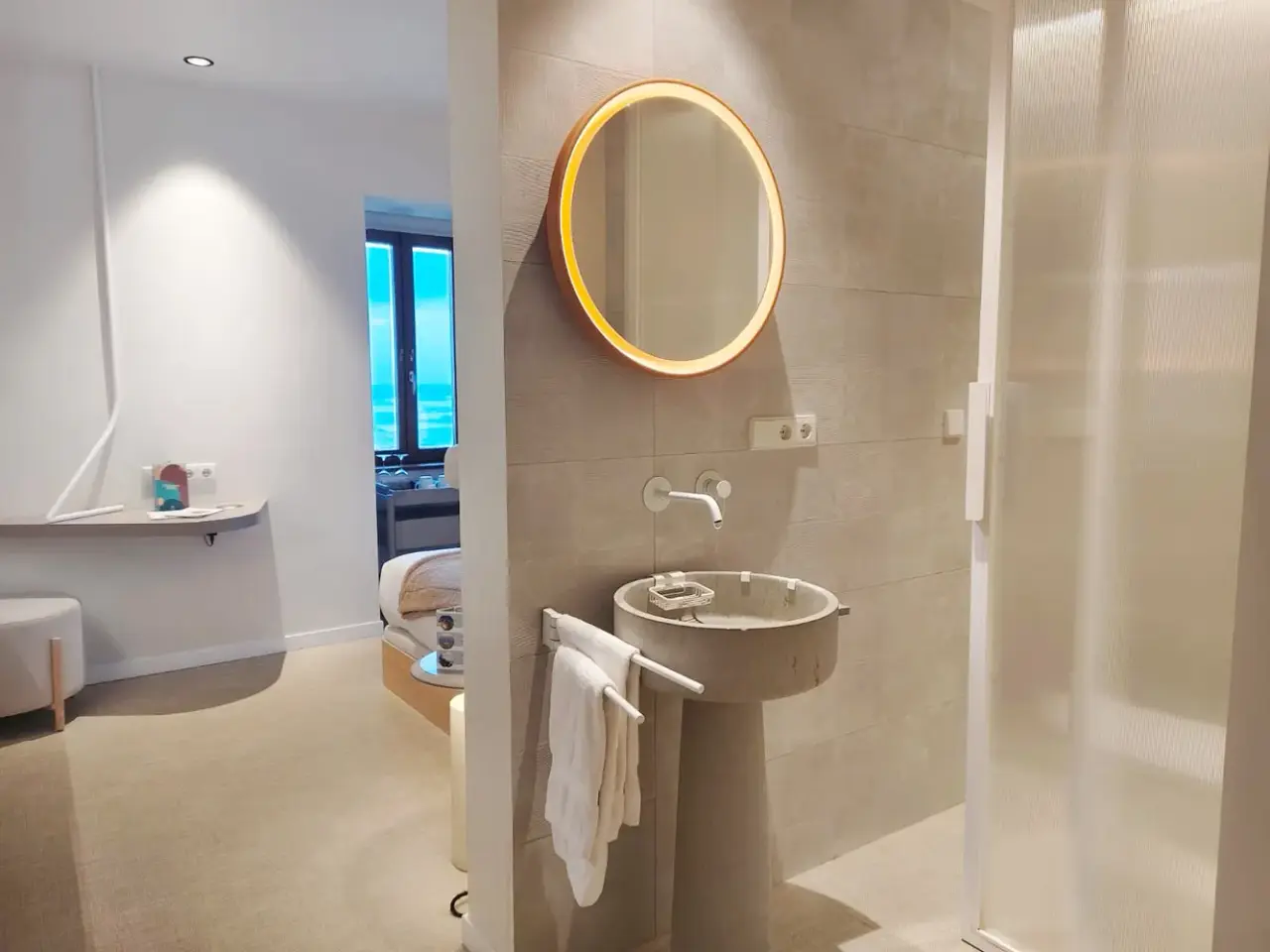 Vista de un baño moderno con lavabo de pedestal circular, grifo mural, espejo redondo retroiluminado y toallas blancas
